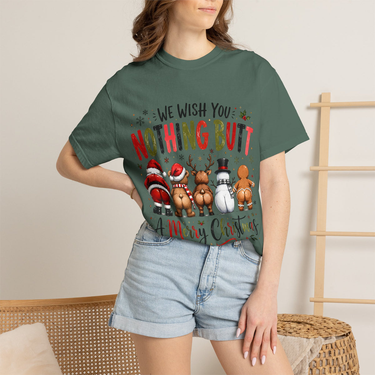 A woman wearing a dark green graphic t-shirt with a Christmas-themed design and light-wash denim shorts, standing in a neutral indoor setting.