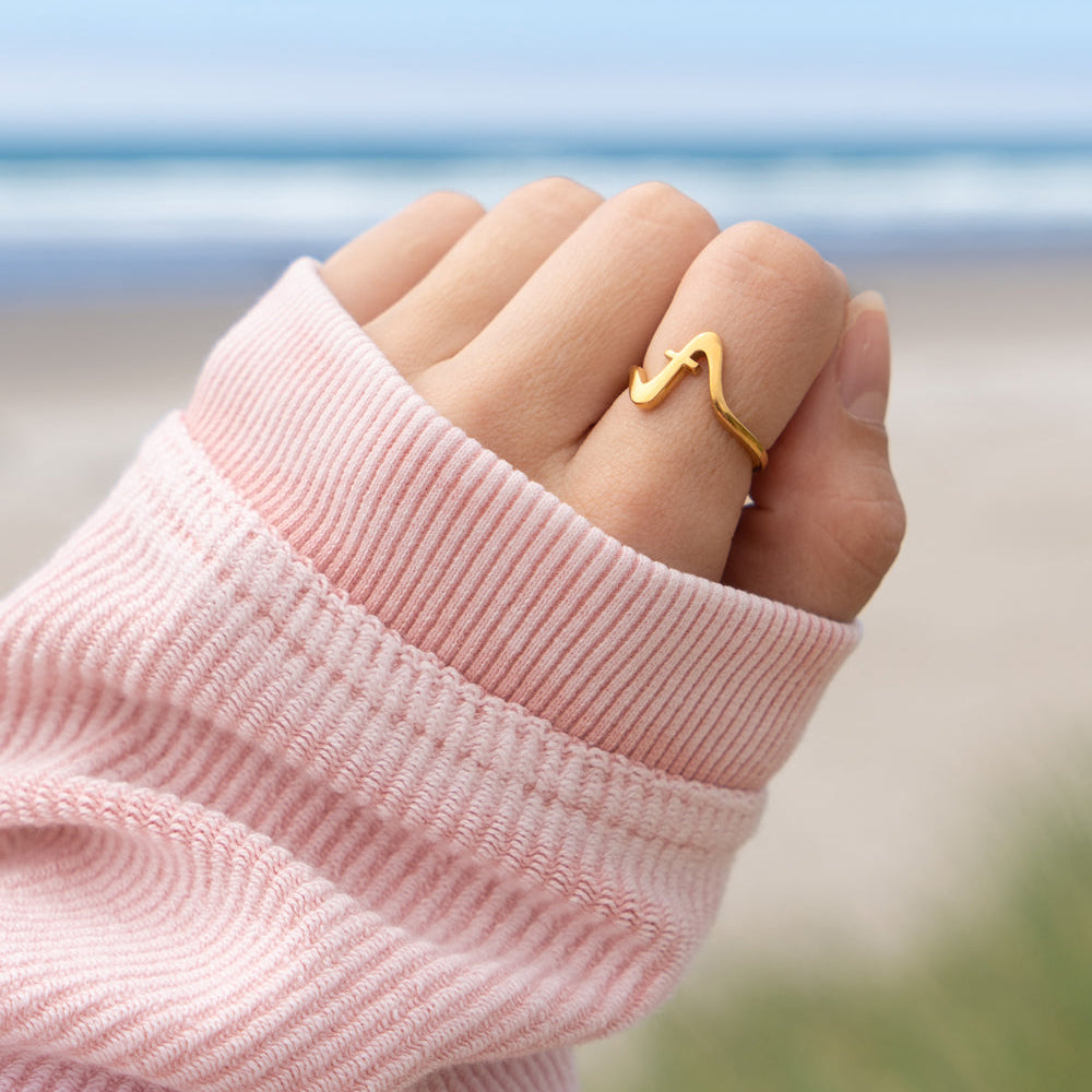 Hand wearing a gold ring with a pink sleeve against a blurred beach background