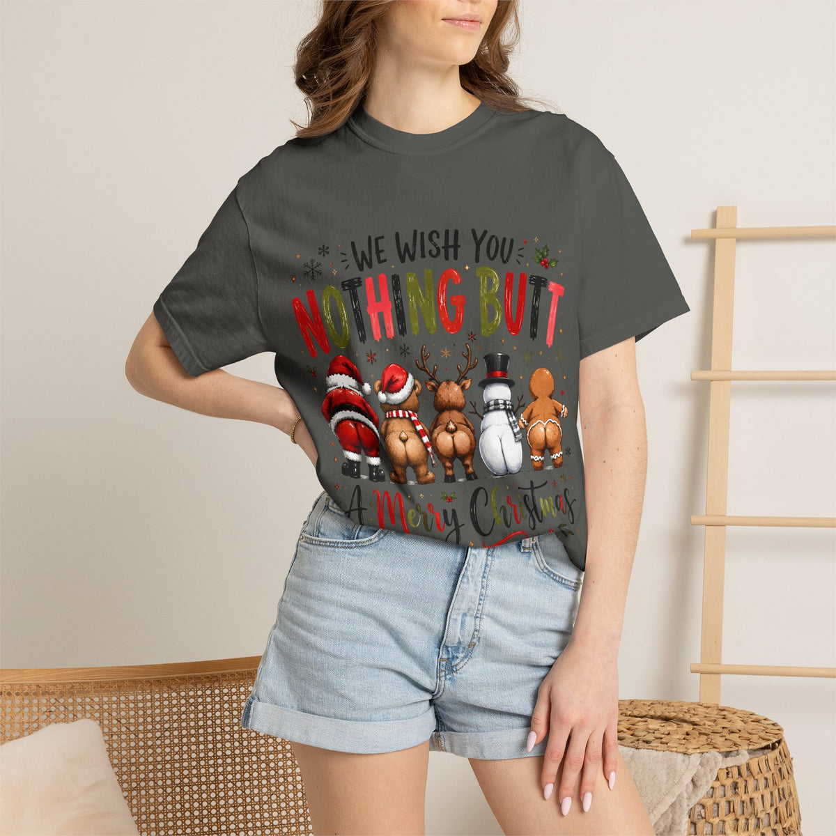 A woman wearing a dark charcoal gray graphic t-shirt with a Christmas-themed 'nothing but merry chaos' print, paired with light-wash denim shorts in a cozy indoor setting.
