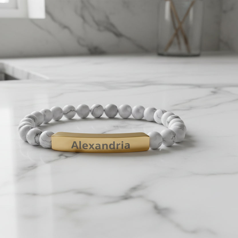 Bracelet with white beads and gold tag displaying 'Alexandria' on a white background