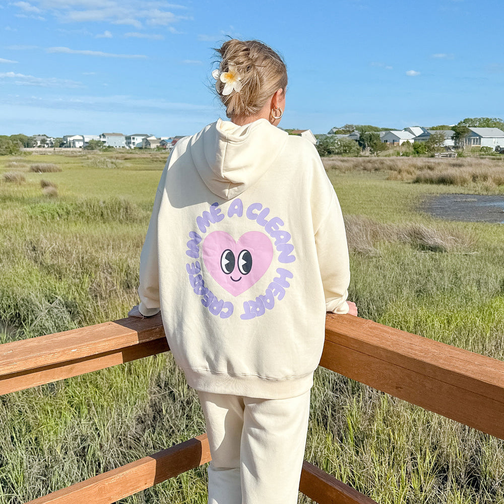 Person wearing a cream-colored hoodie with a graphic design, standing on a wooden platform with a natural landscape in the background.