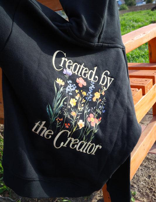 Black hoodie with floral design and text 'Created by the Creator' on a wooden bench.