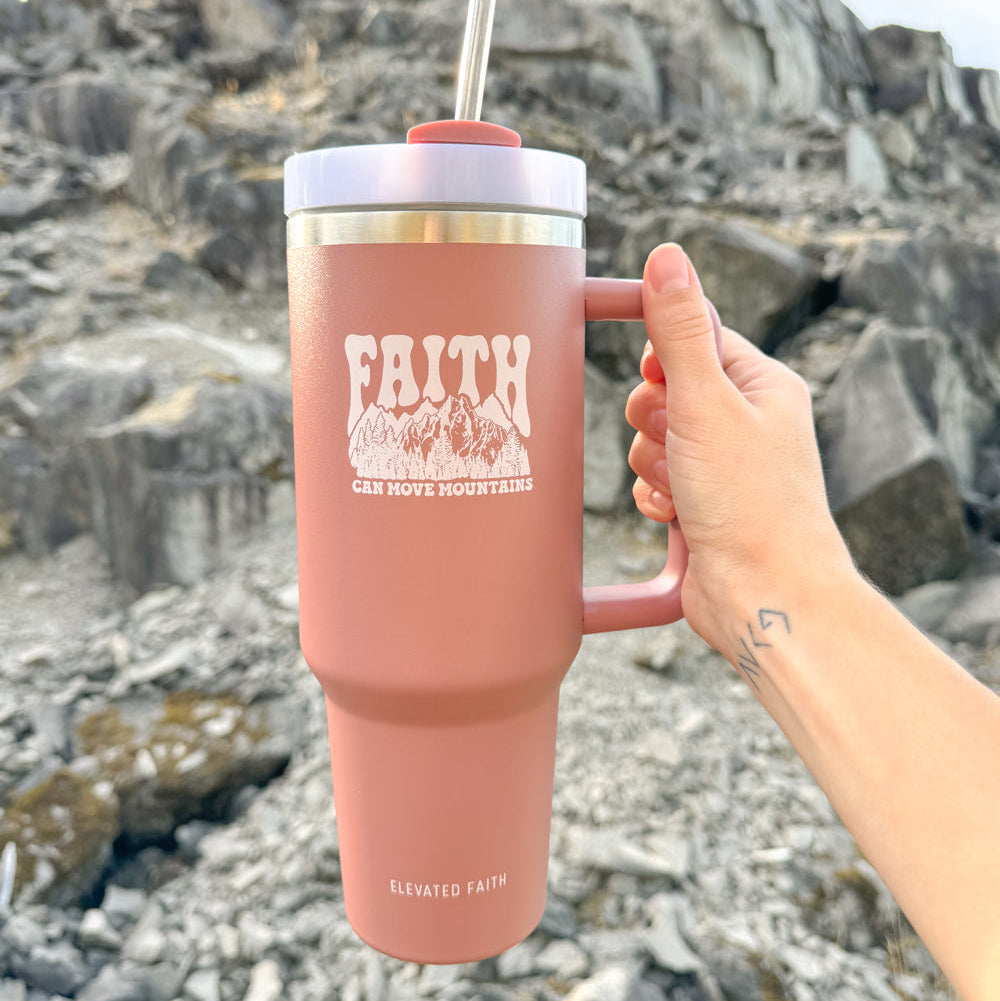 Person holding a pink tumbler with 'Faith Can Move Mountains' text against a rocky background