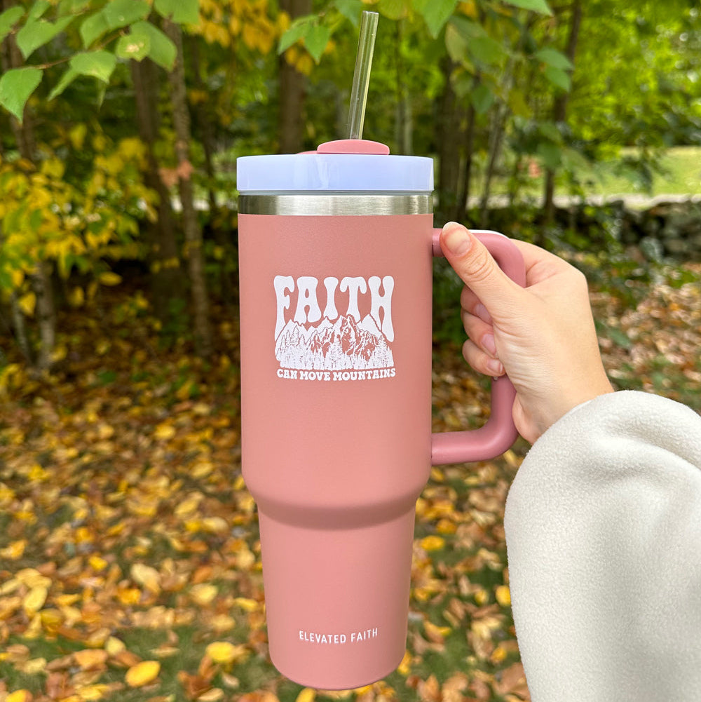 Pink tumbler with 'Faith Can Move Mountains' text held outdoors with trees and fallen leaves in the background