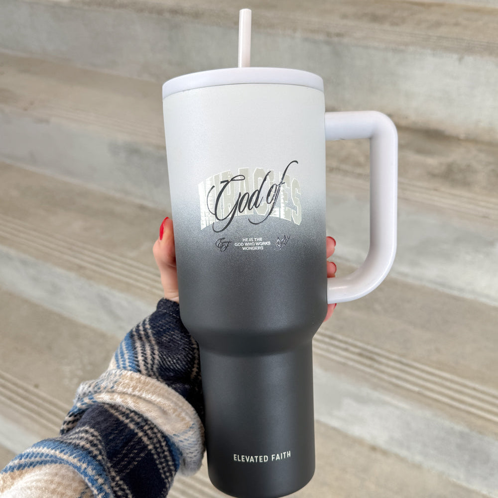 Person holding a black tumbler with a white handle and straw, featuring text on a wooden surface.