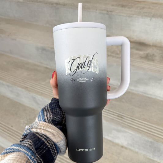 Person holding a black tumbler with a white handle and straw, featuring text on a wooden surface.