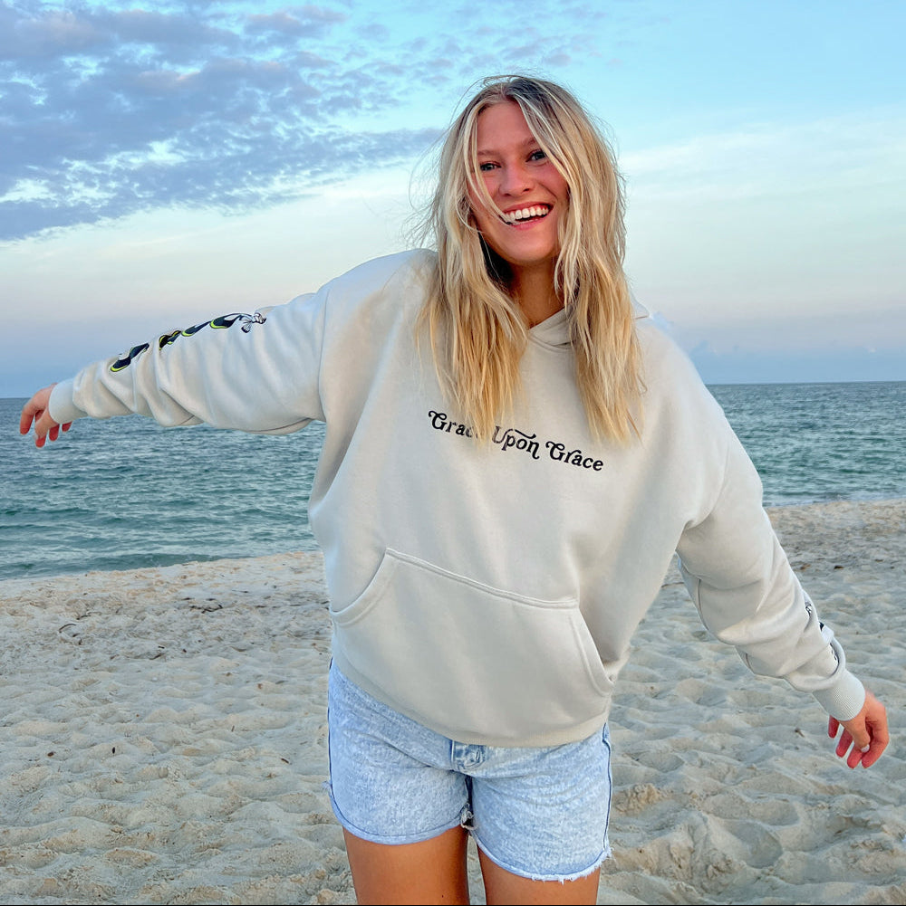 Person wearing a hoodie with text on it, standing on a beach with ocean and sky in the background