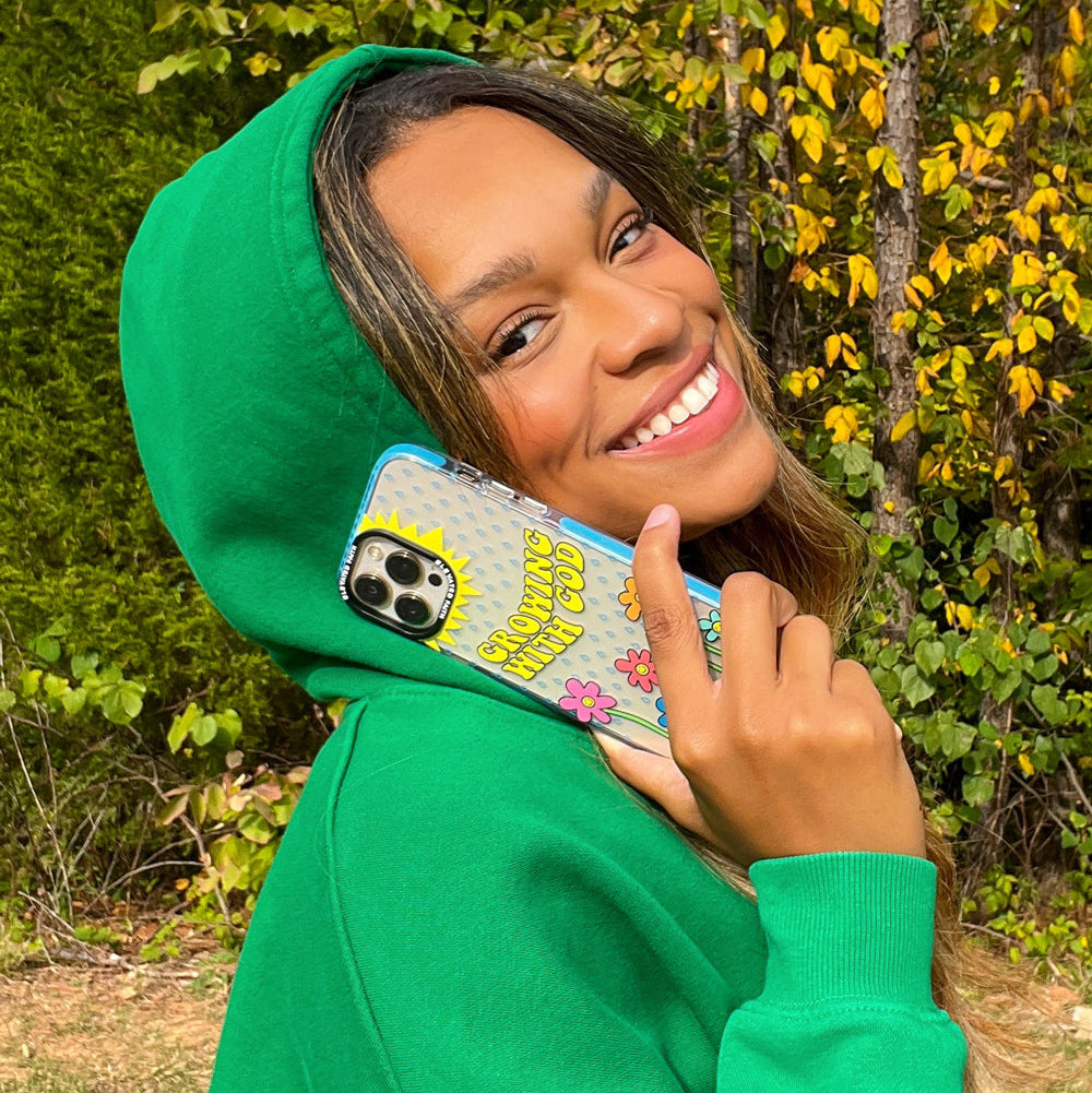 Person wearing a green hoodie holding a phone with a colorful case outdoors.