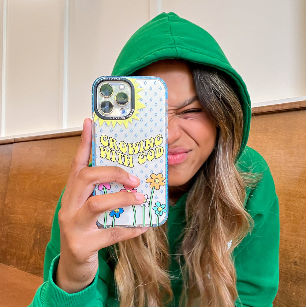 Person wearing a green hoodie holding a phone with a 'Growing with God' case.