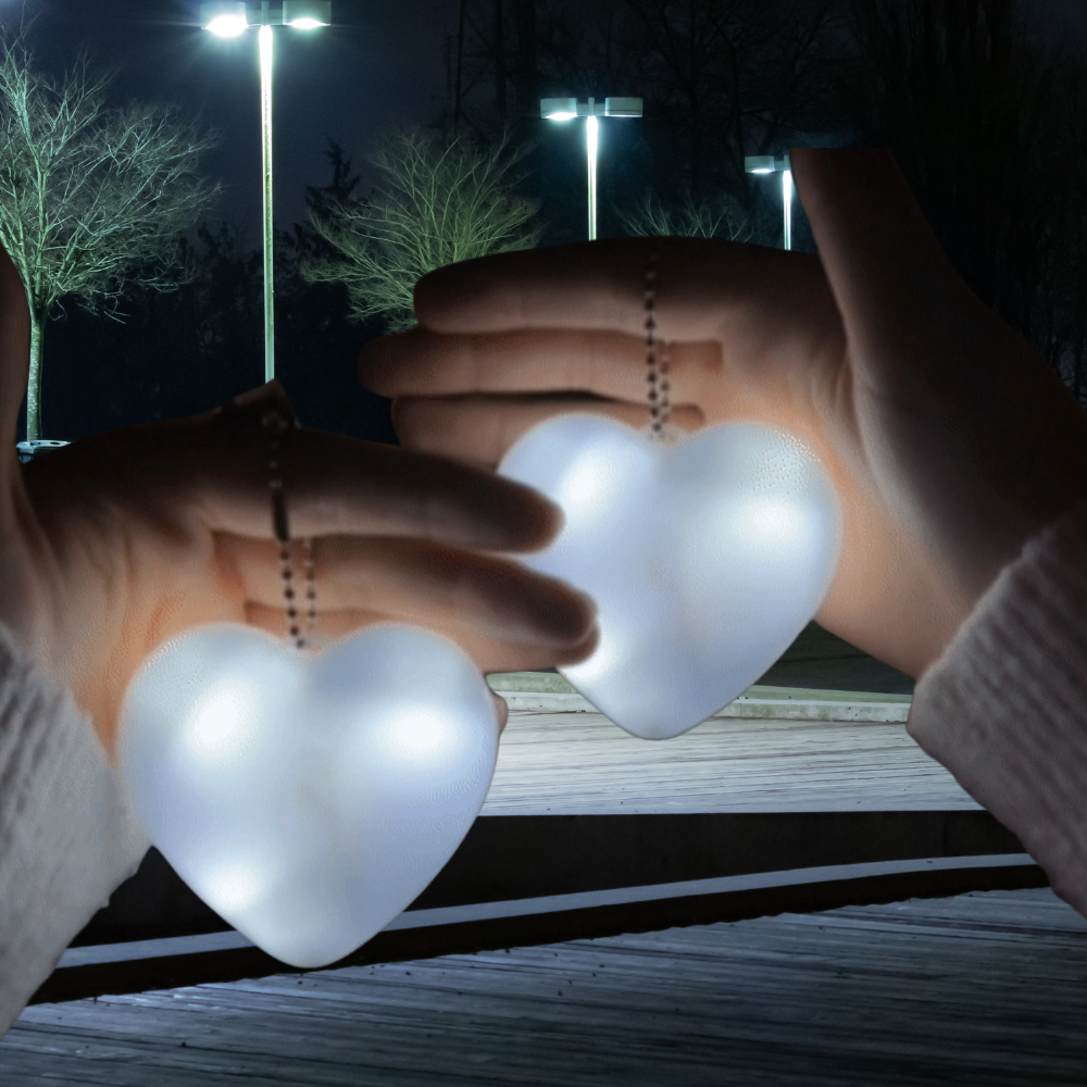Two hands holding glowing heart-shaped pendant lights at night on a wooden walkway, with streetlights and trees blurred in the background.