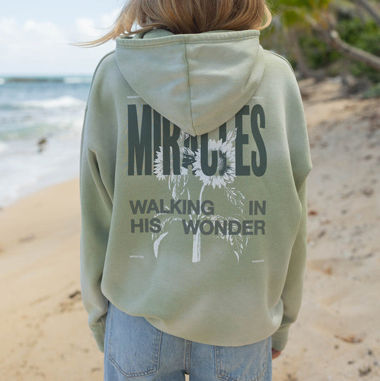 Person wearing a light green hoodie with text on the back, standing on a beach.