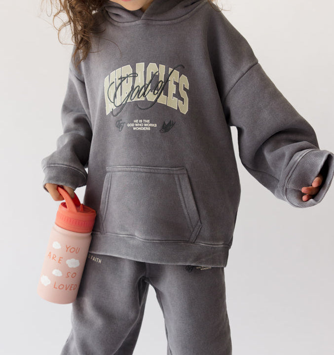 Child wearing a gray sweatshirt and pants set with text, holding a pink water bottle on a white background