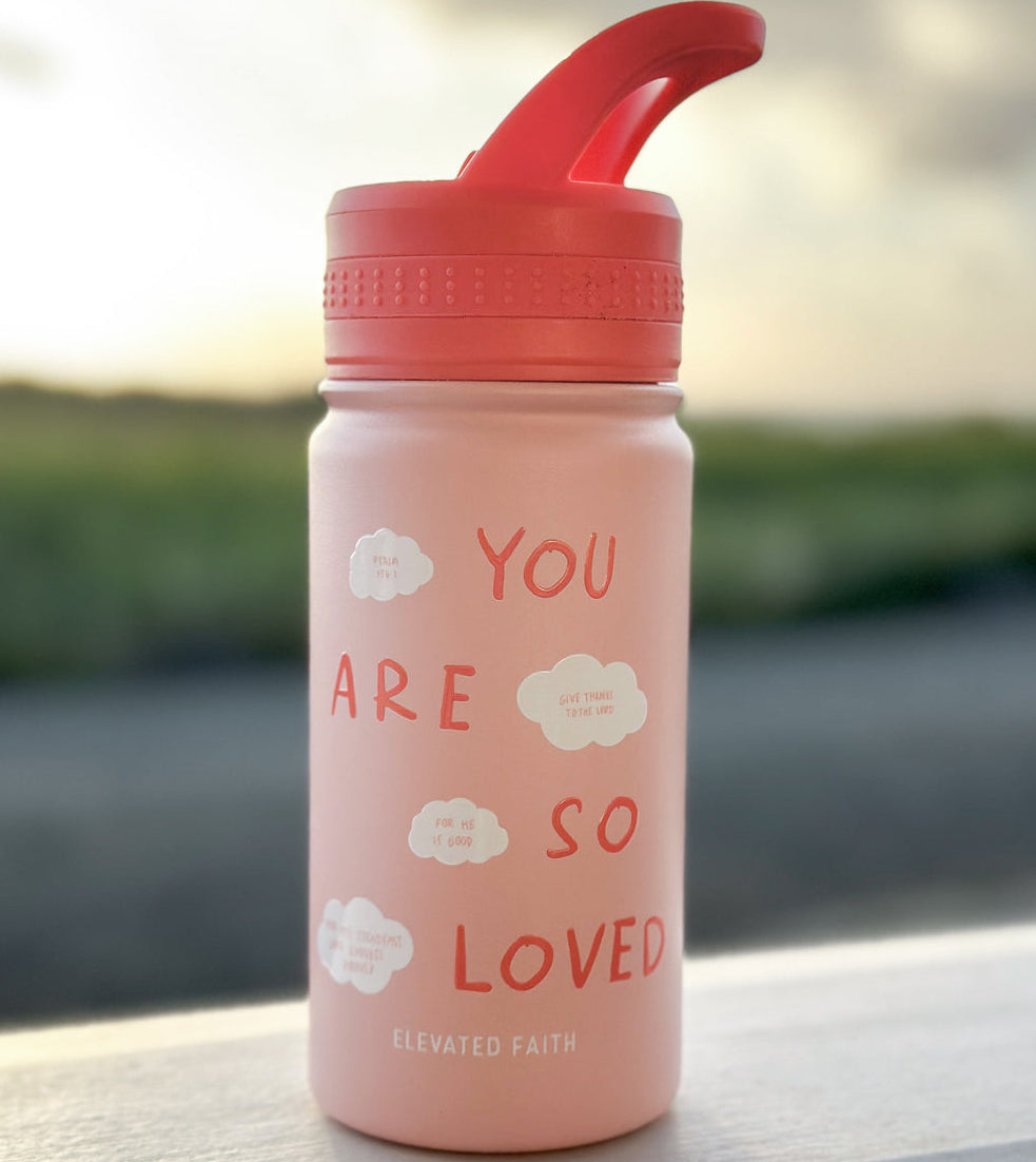 Pink water bottle with red lid and 'You are so loved' text on a blurred natural background